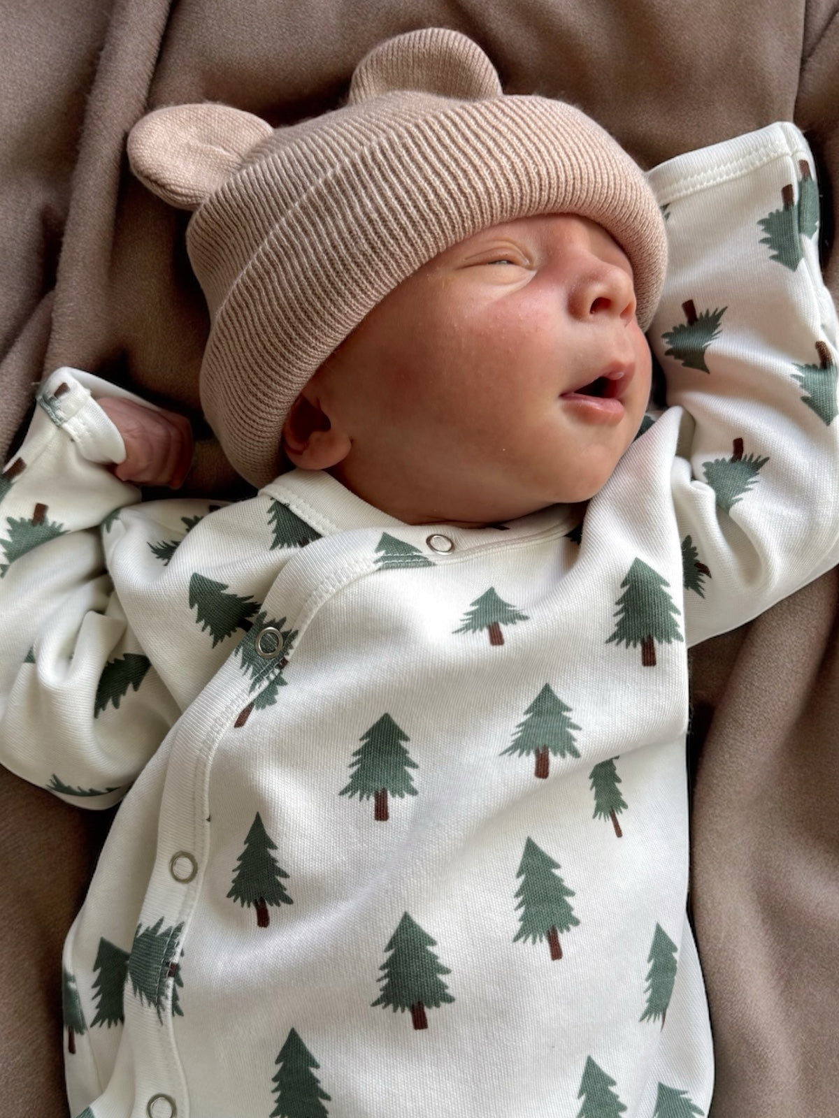 Baby asleep on a soft blanket, wearing a bear-ear hat and a green-tree patterned onesie.