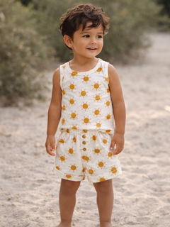 Smiling child in sunny-patterned outfit standing on sandy beach with greenery in the background.