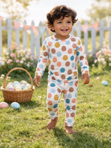 Happy toddler in colorful polka dot pajamas walking on grass, with a basket of pastel eggs nearby and a festive background.