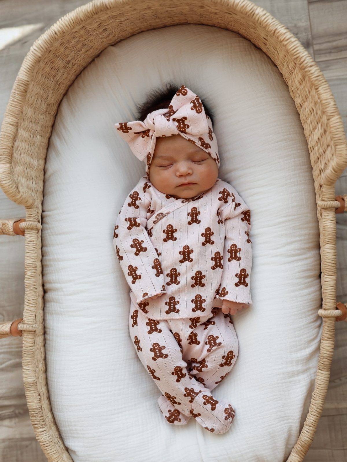 Newborn baby girl sleeping in a wicker bassinet, wearing a gingerbread-patterned outfit with a matching headband.