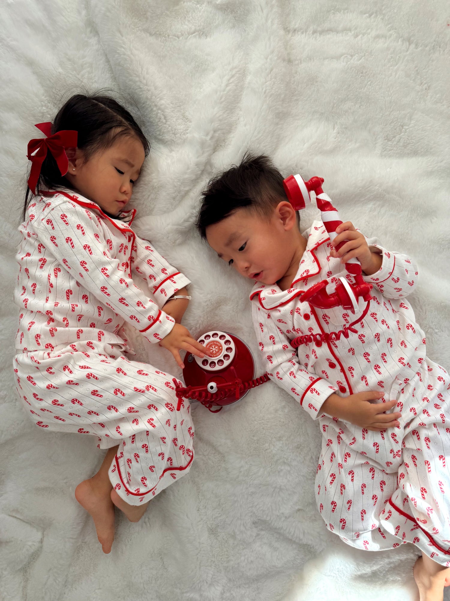 Two children in festive pajamas play with a red rotary phone on a soft white blanket.