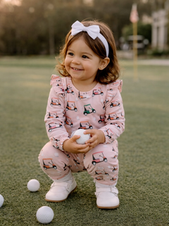 Smiling toddler in pink golf cart pajamas holds a golf ball on a green lawn, with sunshine in the background.