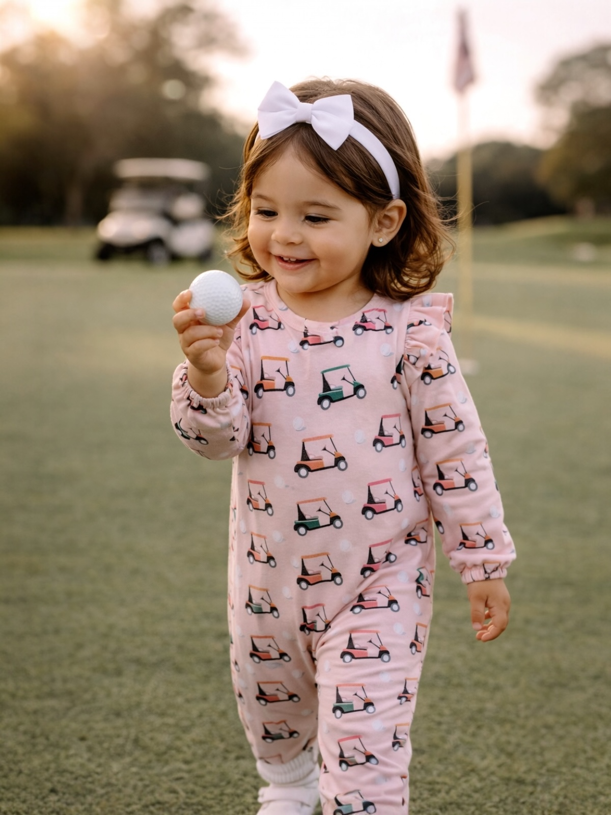 Smiling child in a pink golf-themed outfit holds a golf ball on a course during sunset.