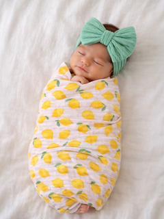 Newborn baby swaddled in lemon-patterned blanket, wearing a mint green bow headband, peacefully sleeping.