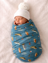 Newborn wrapped in a fish-patterned blanket wearing a white knit hat, sleeping peacefully on a soft surface.