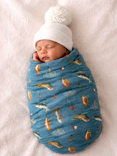 Newborn wrapped in a fish-patterned blanket wearing a white knit hat, sleeping peacefully on a soft surface.