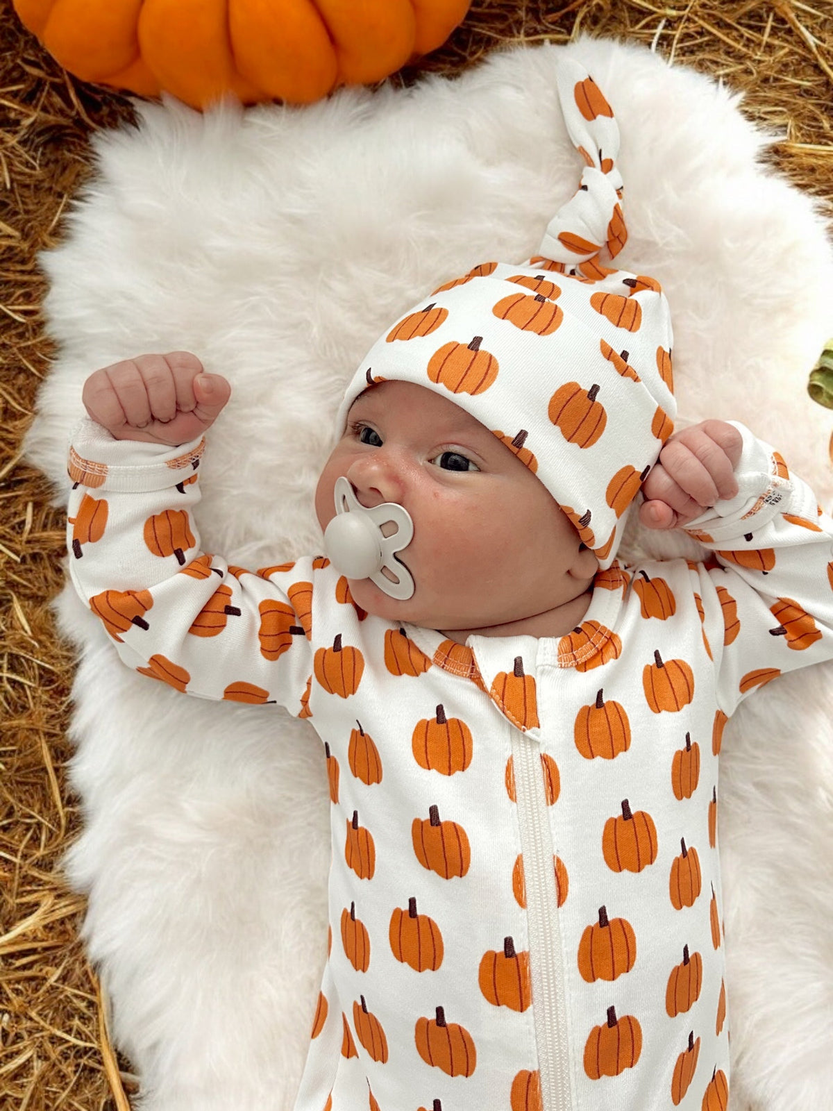Baby in pumpkin-patterned outfit lying on a soft surface, with a pumpkin in the background.