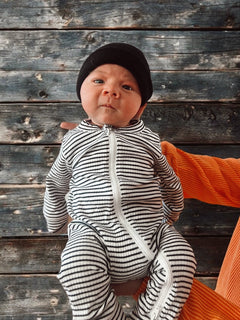 Infant in a striped onesie and black beanie held against a wooden background.