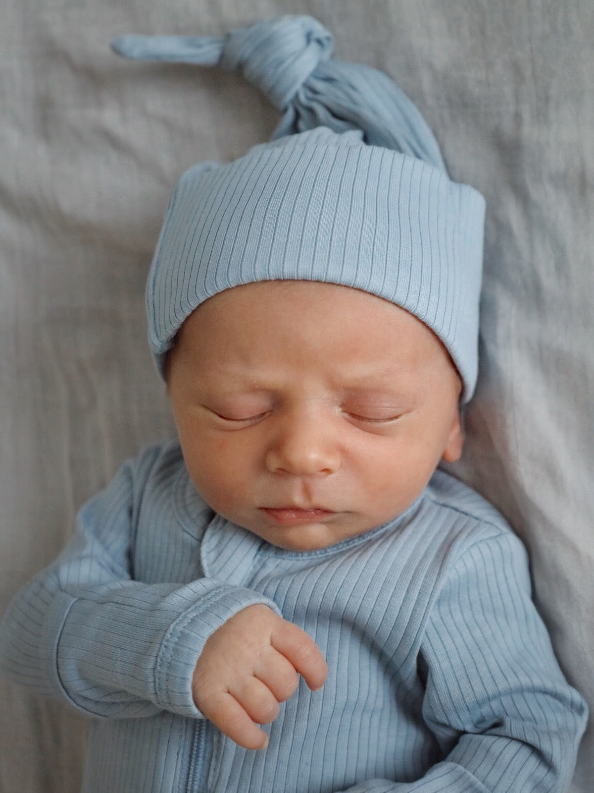 Newborn baby peacefully asleep in a light blue outfit with a matching hat, resting on a soft, neutral background.