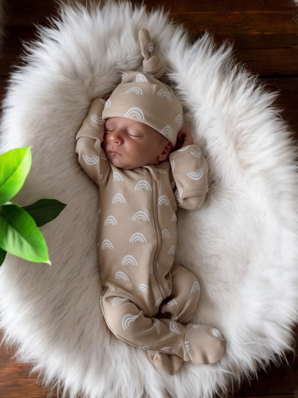 Newborn sleeping on a soft faux fur blanket, wearing a beige outfit with white rainbow patterns and a matching hat. 