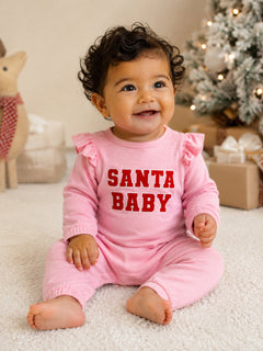 Smiling baby in pink "Santa Baby" outfit, seated on a soft rug with holiday decorations in the background.