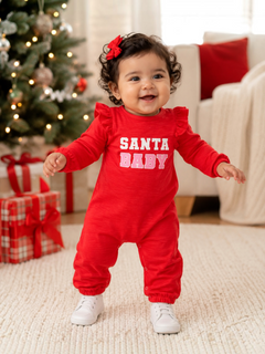 Smiling baby in red "SANTA BABY" outfit stands near a decorated Christmas tree with presents.