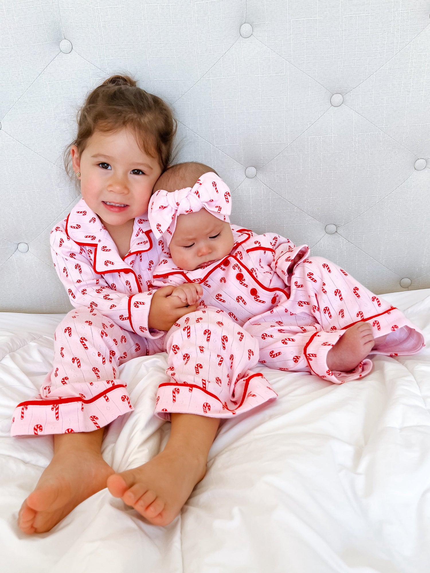 Two young girls in matching pink candy cane pajamas sit on a bed, smiling and cuddling together.