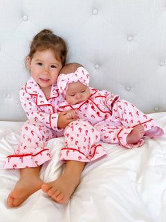 Two young girls in matching pink candy cane pajamas sit on a bed, smiling and cuddling together.