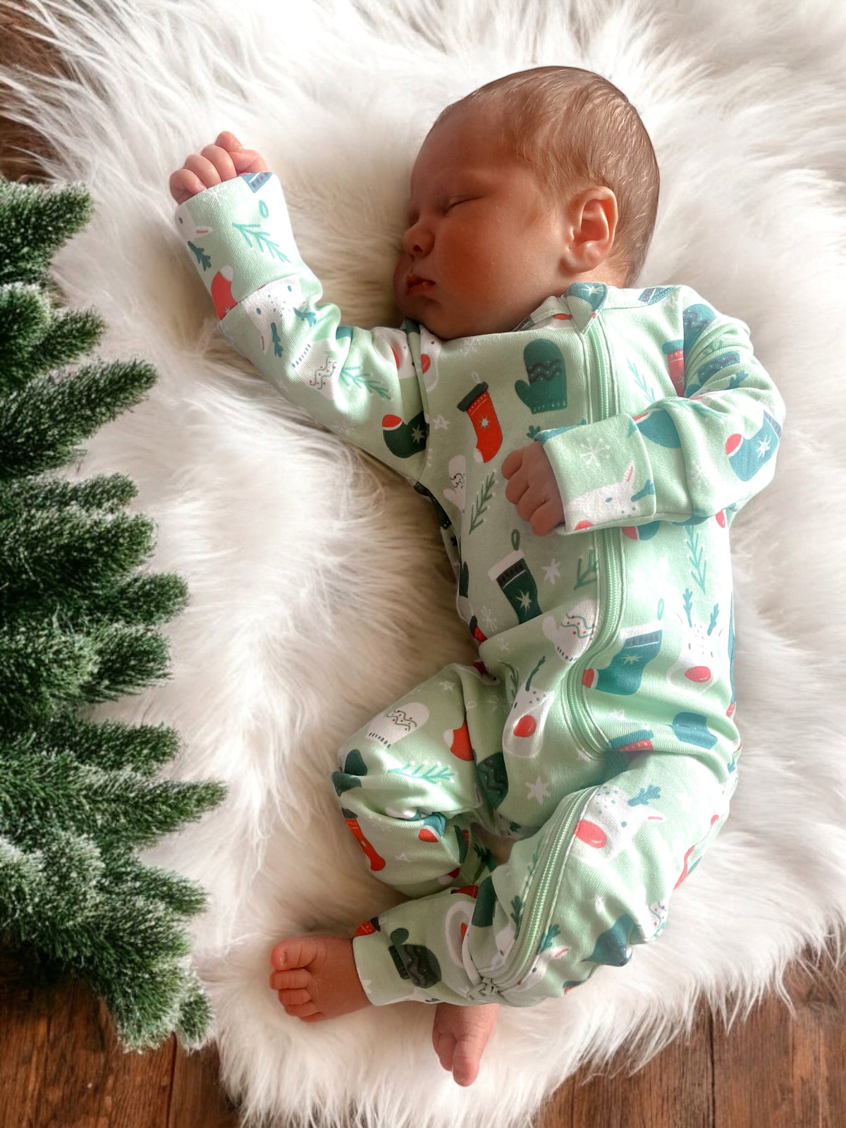 Sleeping baby in festive green pajamas on soft white fur rug, surrounded by pine tree branches.