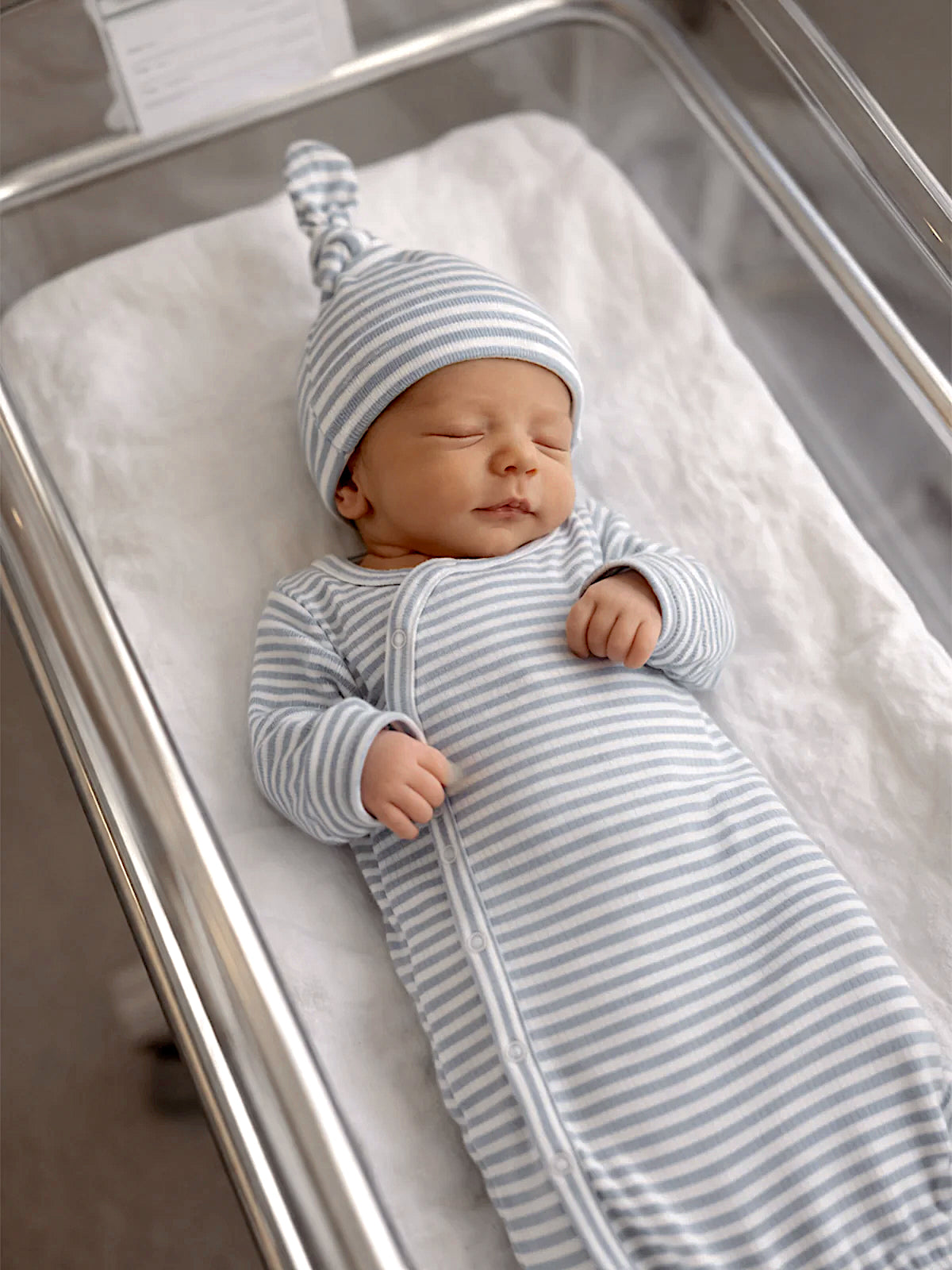 Newborn baby peacefully sleeping in a striped outfit and hat in a hospital bassinet.