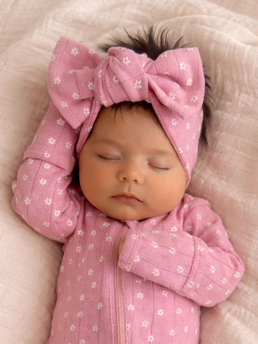 Sleeping baby girl in a pink outfit and large bow on a soft blanket.