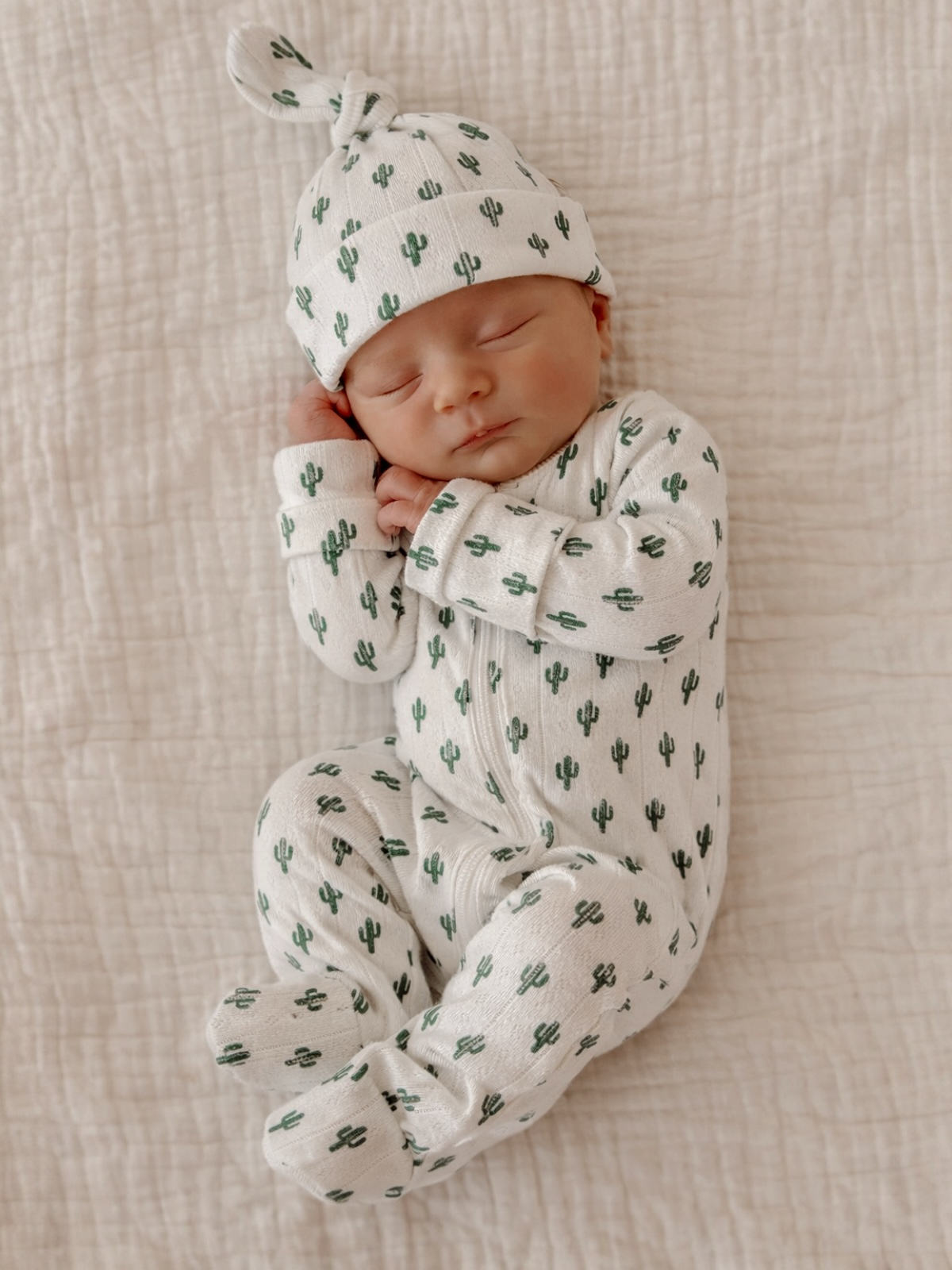 Newborn baby sleeping peacefully in cactus-patterned outfit and hat on a textured white blanket.