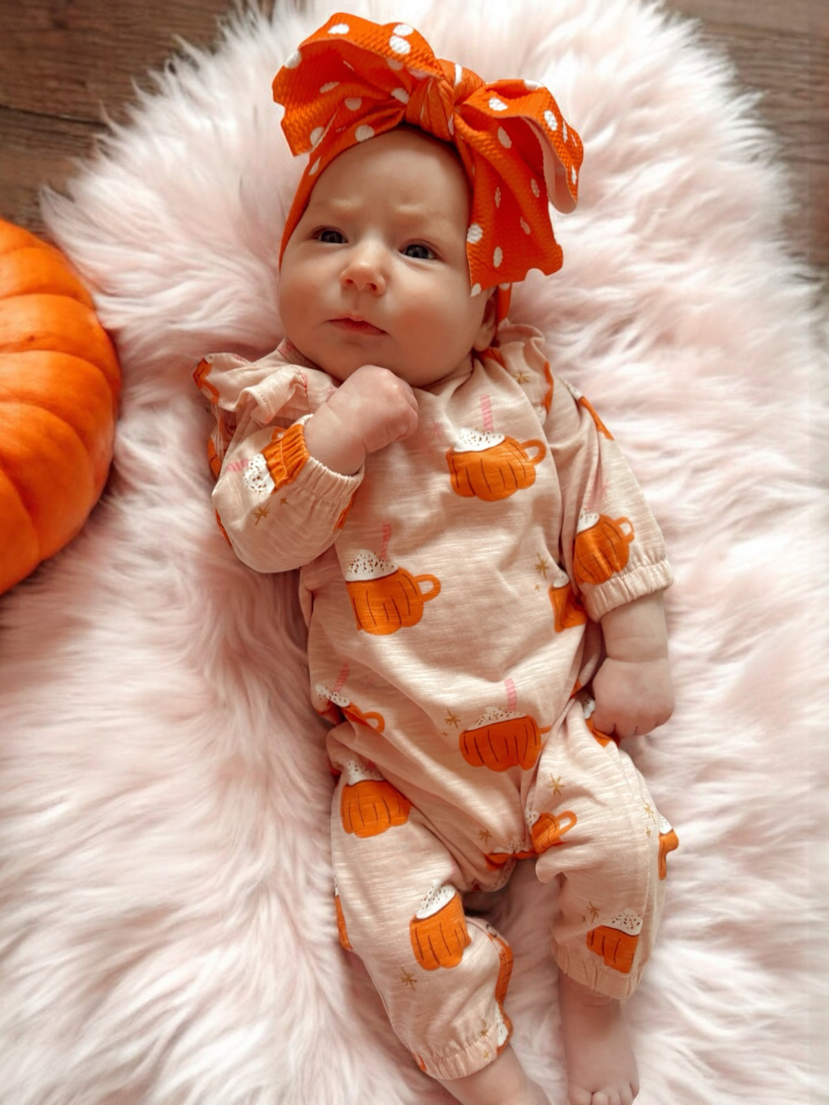 Baby girl in cute pumpkin-themed outfit and large orange polka dot bow, lying on soft pink blanket with a pumpkin nearby.