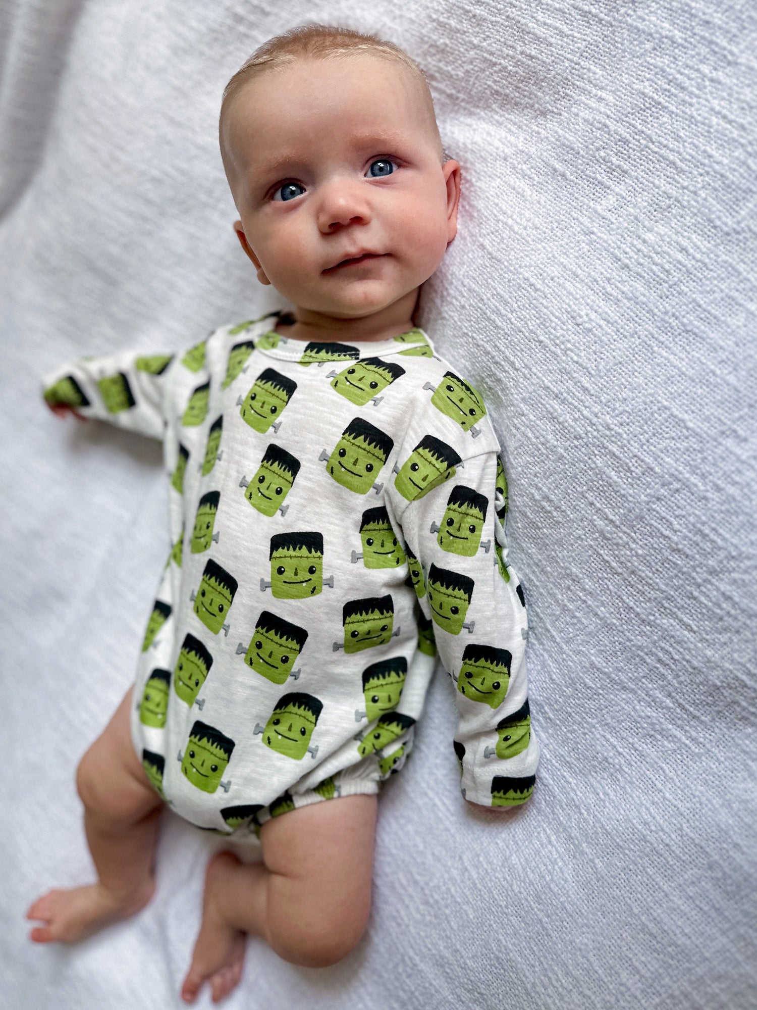 Baby in a Frankensteins-patterned onesie, looking curiously at the camera on a textured white surface.
