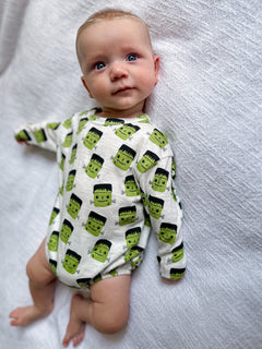 Baby in a Frankensteins-patterned onesie, looking curiously at the camera on a textured white surface.