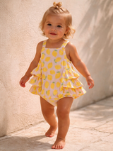 Smiling toddler in a lemon-patterned romper walking on a stone pathway. Natural light and neutral background.