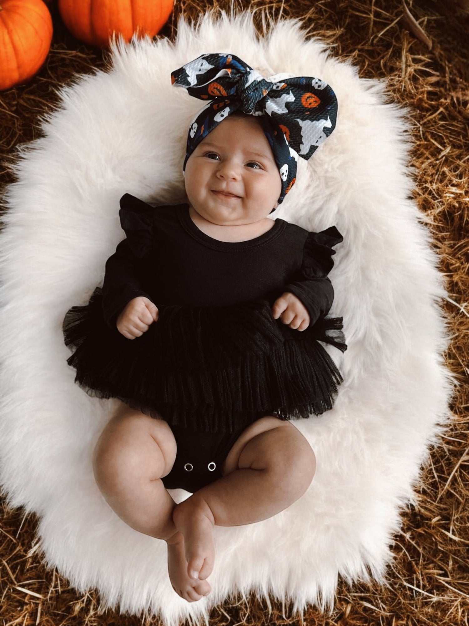 Baby girl in a black dress and ghost-patterned bow headband, smiling on a fluffy white blanket with pumpkins around.