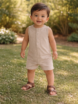 Smiling toddler in a beige romper and brown sandals, standing on green grass with soft sunlight filtering through trees.