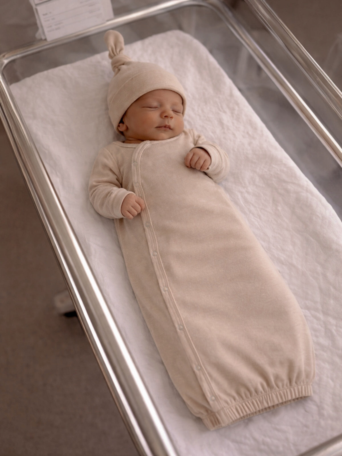 Sleeping newborn in a beige swaddle and knit hat, resting peacefully in a hospital bassinet.