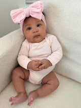Baby girl sitting on a plush chair, wearing a pink headband and striped onesie, with hands clasped in front.