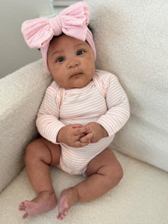 Baby girl sitting on a plush chair, wearing a pink headband and striped onesie, with hands clasped in front.