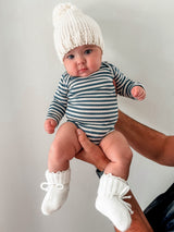 Baby in a white knitted hat and striped outfit, being held by a person against a light background.