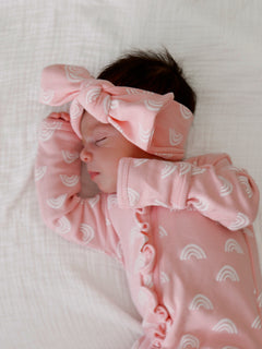 Sleeping baby in a pink outfit with rainbow patterns and a matching headband on a soft white blanket.