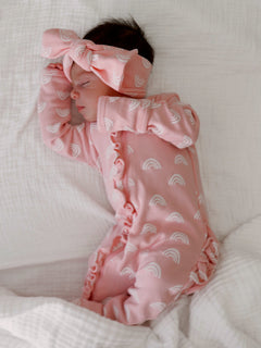 Sleeping baby in a pink rainbow-patterned onesie and matching headband, nestled in soft white blankets.
