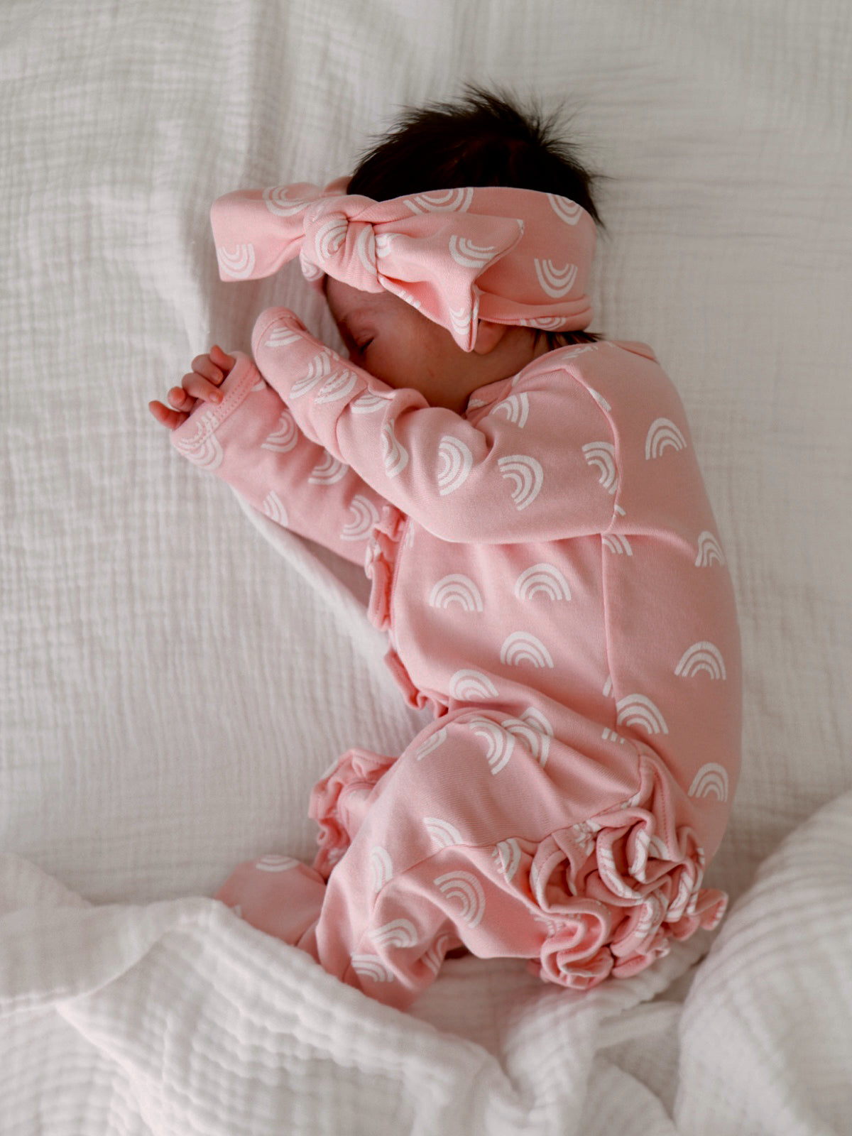 Infant sleeping peacefully in pink pajamas with rainbow patterns, wearing a matching headband on a white blanket.