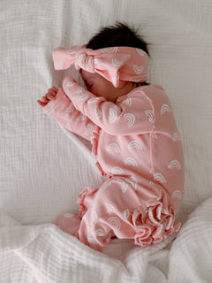 Infant sleeping peacefully in pink pajamas with rainbow patterns, wearing a matching headband on a white blanket.