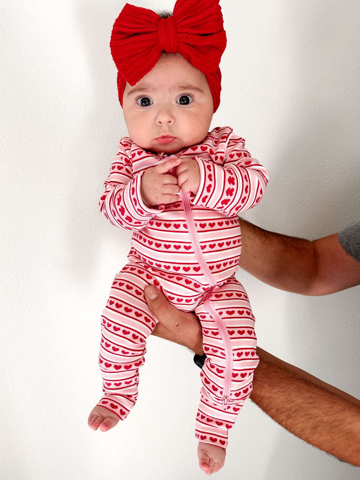 Baby in a red heart-patterned onesie and large red bow, held by a hand against a white background.