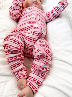 Baby in a pink striped onesie with red hearts, lying on a white blanket, raising arms playfully.