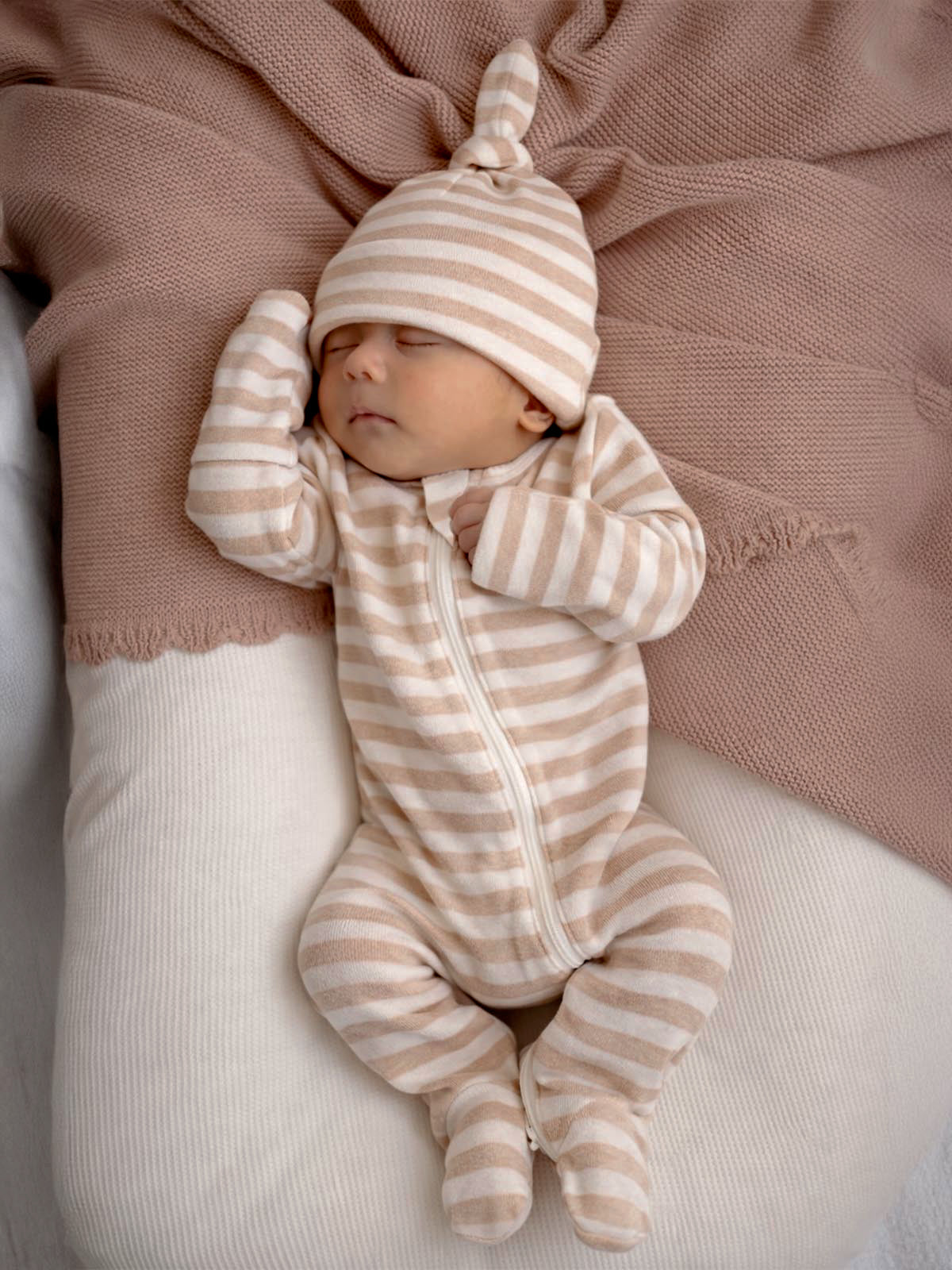 Baby sleeping peacefully in striped pajamas and hat, surrounded by soft, textured fabrics in neutral tones.