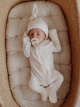 Baby in striped onesie and hat sleeps in a cozy basket, holding a pacifier on a cushioned surface.