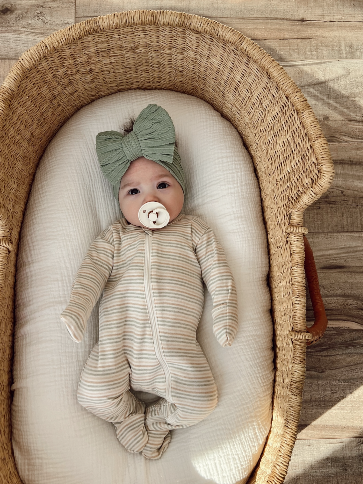 Infant in a striped onesie and green bow headband resting in a woven bassinet with a pacifier.