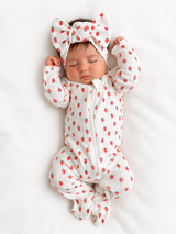 Sleeping baby in a strawberry-patterned onesie and bow headband, resting on a white blanket.