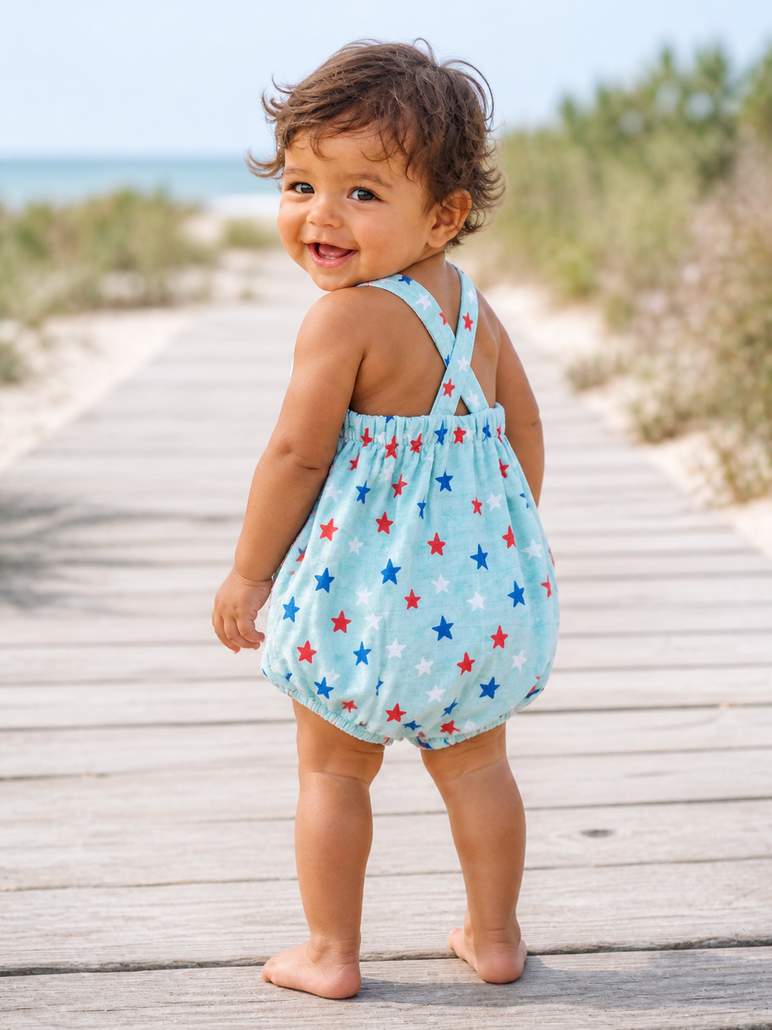 Smiling toddler in a star-patterned outfit, standing on a wooden path by the beach.