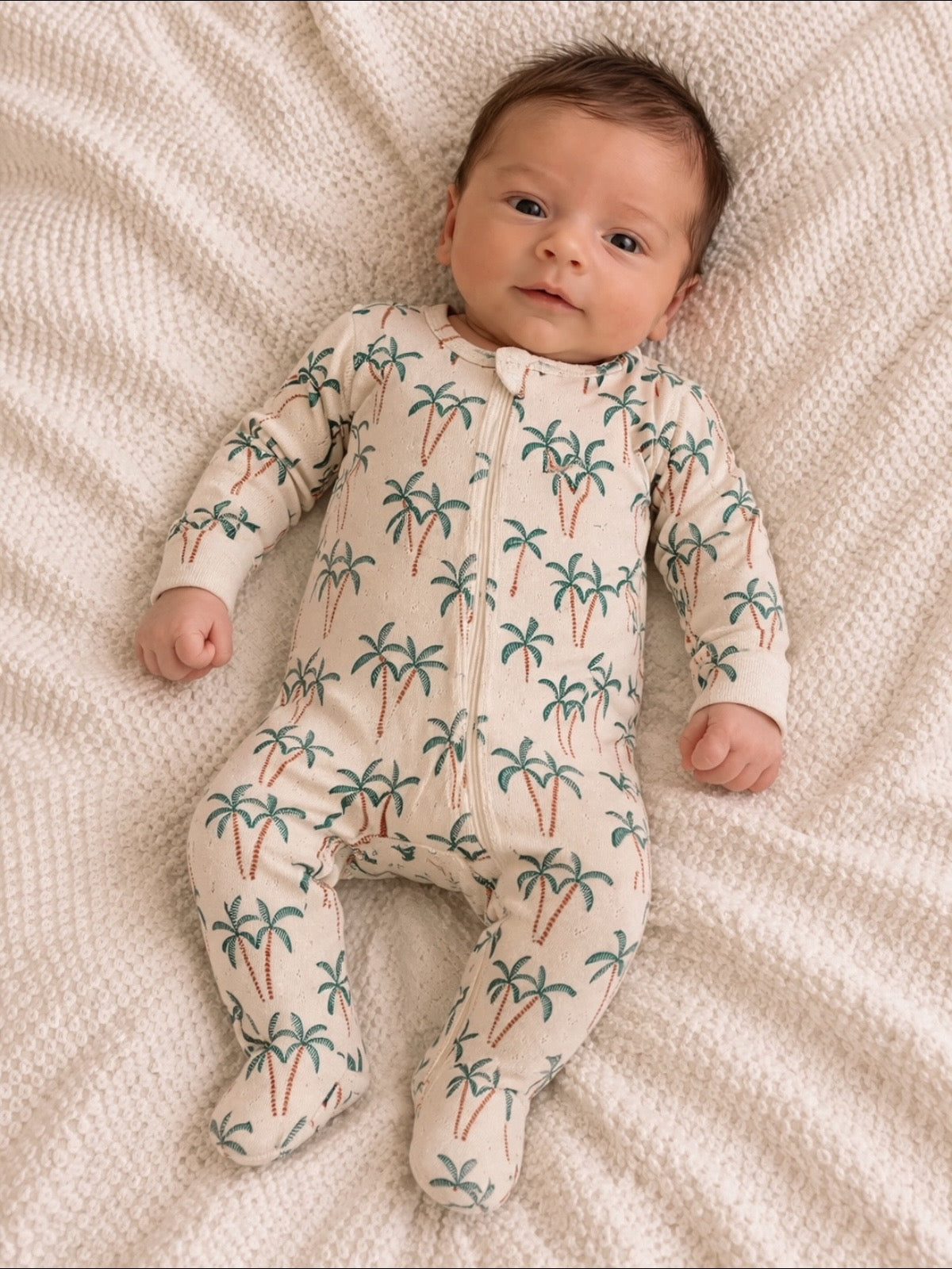 Baby in palm-print onesie lying on a textured white blanket, looking curiously at the camera.