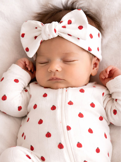 Sleeping baby wearing a white outfit with red dots and a large bow headband, lying on a soft surface.