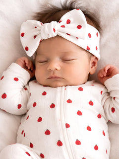 A sleeping baby in a polka dot onesie with a large bow headband, resting on a soft white surface.