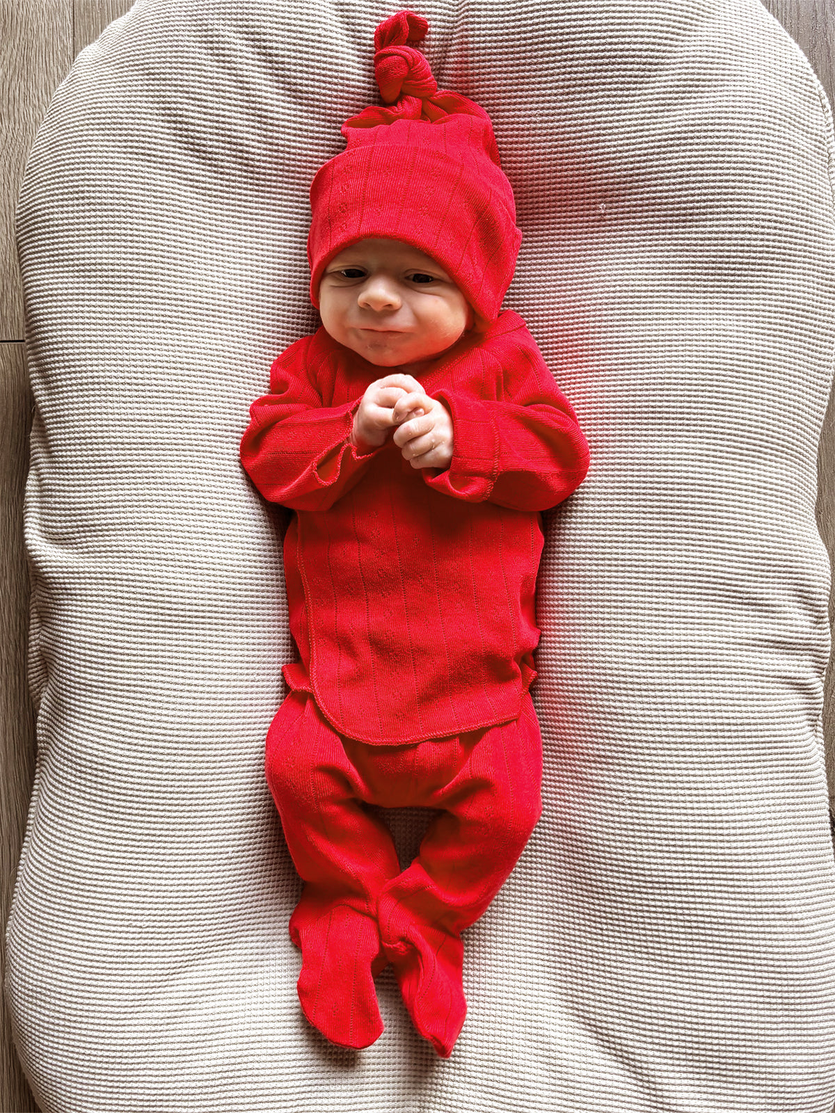 Baby in a red outfit and hat, smiling while lying on a textured cream-colored surface.