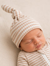 Sleeping newborn wearing a striped hat and sleep suit, resting peacefully on a soft, neutral-colored surface.