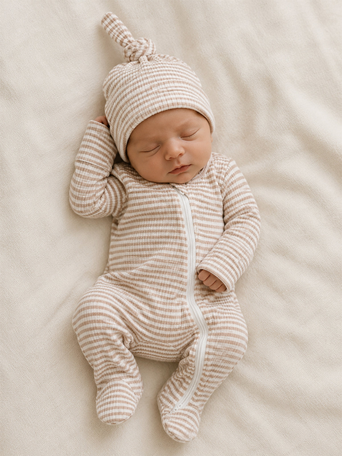 Newborn baby in a striped onesie and matching hat, peacefully sleeping on a soft, light-colored blanket.