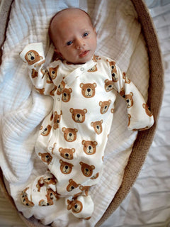 Baby in a bear-patterned onesie, lying in a woven basket with a soft white blanket. Gentle, curious expression.